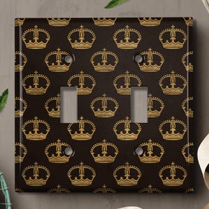 May include: A black light switch cover with a gold crown pattern. The cover has two toggle switches.