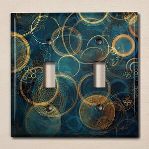 May include: A decorative double light switch cover with an abstract design. The cover has a dark teal background with overlapping gold and teal circles and lines. The light switches are silver.