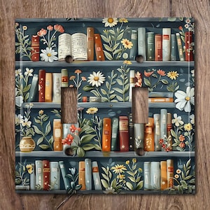 May include: A light switch cover with a whimsical design featuring a bookshelf filled with books, flowers, and greenery. The bookshelf is painted in a dark blue colour with a light blue background.