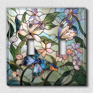 May include: A decorative double light switch cover featuring a stained glass design. The cover showcases a vibrant floral and butterfly motif with shades of blue, green, pink, and cream. The butterflies have intricate wing patterns.