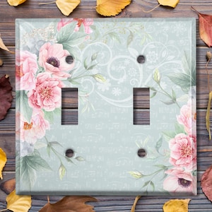 May include: A light blue double light switch cover with a floral design. The design features pink and white flowers with green leaves on a light blue background with a white musical note pattern.