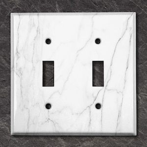 May include: A white marble-patterned double light switch cover with two rectangular switch openings and four screw holes. The cover has a glossy finish and a square shape. The marble design features gray veining.
