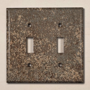 May include: A double light switch plate with a rustic, brown and grey mottled finish. The plate has a rectangular shape with rounded corners.