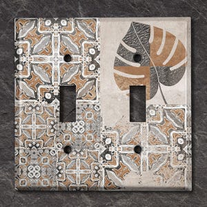 Puede incluir: Una tapa de interruptor de luz con un diseño decorativo que presenta un patrón de hojas marrón y gris y un patrón de azulejos geométricos. La tapa del interruptor está hecha de cerámica y tiene un fondo blanco.