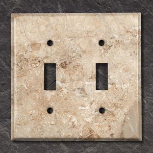 May include: A double light switch cover with a beige marble pattern. The cover has two toggle switches and two screw holes.