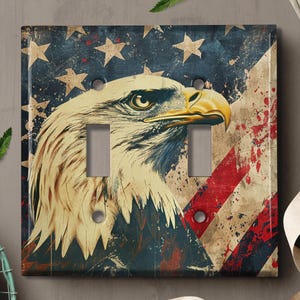 May include: A light switch cover featuring a distressed American flag with a bald eagle in the foreground. The eagle's head is facing the right side of the image. The eagle's eye is visible. The eagle's beak is open. The eagle's feathers are detailed. The flag is faded and has red, white, and blue colors. The flag has a distressed look with paint splatters.