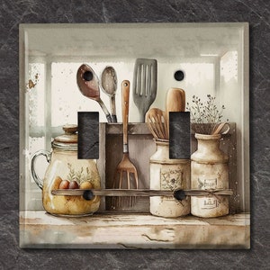 May include: A light brown double light switch cover featuring a watercolour painting of a kitchen shelf with jars, utensils, and dried flowers. The shelf is painted in a light brown colour with a white background.