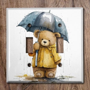 May include: A watercolour illustration of a brown teddy bear wearing a yellow raincoat and holding a blue umbrella. The bear is standing in a puddle of water with raindrops falling around it.