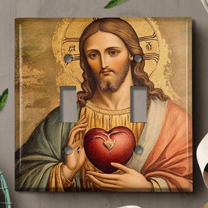 May include: A light switch cover featuring a painting of Jesus Christ with a heart in his hand. The painting is in a traditional style with a gold background and ornate details.