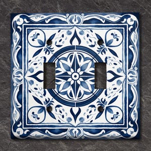 May include: A white light switch cover with a blue and white patterned design. The design features a central motif of a starburst with floral accents.