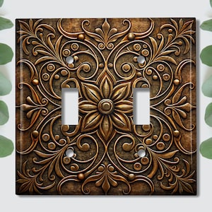May include: Ornate bronze-colored double light switch cover with a floral and scroll design. The cover features a central flower motif and symmetrical patterns. The background has a textured, aged appearance.