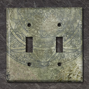 May include: A decorative double light switch cover with a detailed mandala design. The cover has a distressed, textured appearance with a muted green and gray color palette. The intricate pattern is etched into the surface, adding depth and visual interest.