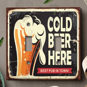 May include: A vintage-style light switch cover with a distressed metal finish. It features a cartoon illustration of a beer mug with the text "COLD BEER HERE BEST PUB IN TOWN!"