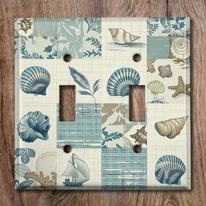 May include: A light blue and white light switch cover with a seashell and nautical themed patchwork design. The design features seashells, a sailboat, and a plaid pattern.