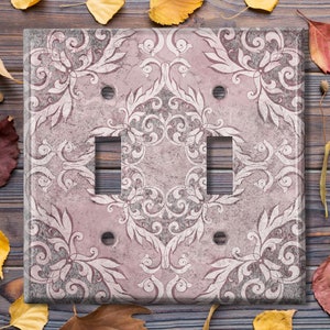 Puede incluir: Una tapa de interruptor de luz con un patrón floral rosa y blanco. La tapa tiene dos interruptores de palanca y una sola toma de corriente.