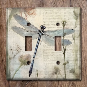 May include: A light blue dragonfly with black markings is centered on a light beige background with a vintage floral pattern and handwritten text. The dragonfly is perched on a stem with a flower bud. The background is a light beige with a vintage floral pattern and handwritten text.