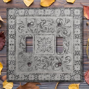 May include: A gray light switch cover with a decorative floral design. The cover has a distressed look and a black border.