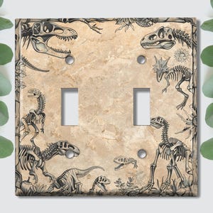 May include: Light brown ceramic light switch cover featuring a dinosaur skeleton design. The cover depicts multiple dinosaur skeletons in various poses, including a T-Rex, a Stegosaurus, and a Triceratops.