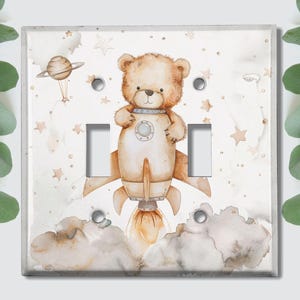 May include: A light brown teddy bear wearing a rocket ship costume with a white background and stars. The rocket ship has a brown and white design with a brown and white cloud behind it.