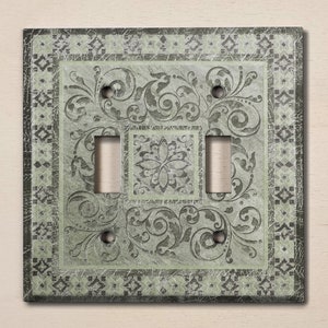 Puede incluir: Una placa de interruptor doble de color verde claro con un patrón floral decorativo y un acabado desgastado.