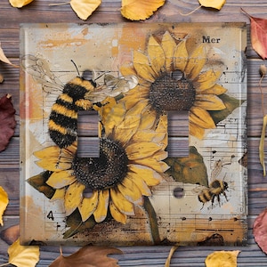 May include: A light switch cover with a vintage design featuring two sunflowers, a bee, and musical notes. The background is a faded, cream-colored paper with a distressed look. The text "Mer" is visible in the upper right corner.