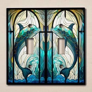 May include: A stained glass light switch cover featuring two blue dolphins leaping through a wave pattern. The dolphins are outlined in gold and the background is a mix of blue, green, and white.