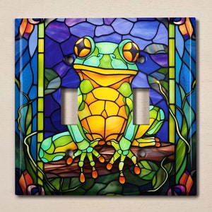 May include: A stained glass light switch cover featuring a green and yellow frog sitting on a branch. The frog has large, round eyes and a wide smile. The background is a vibrant blue with green and purple accents.