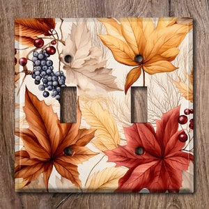 Peut inclure: Une plaque de recouvrement de double interrupteur avec un motif aquarelle de feuilles d'automne dans des tons de rouge, orange, jaune et marron. Les feuilles sont disposées de manière aléatoire sur un fond blanc. On trouve également de petites grappes de baies bleues et rouges dans tout le motif.