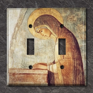 May include: A decorative light switch cover featuring a religious painting. The artwork depicts a figure with a golden halo, wearing a brown robe, and a head covering. The cover has two switch openings and black screw holes.