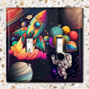 May include: A colorful double light switch cover featuring a rocket ship, planets, and an astronaut in space. The design includes vibrant hues of orange, yellow, blue, and purple, creating a whimsical, space-themed aesthetic.