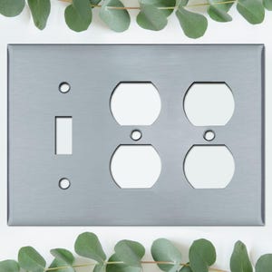 May include: A brushed silver metal wall plate with a toggle switch and four electrical outlet openings. The plate is rectangular with rounded corners. Green eucalyptus leaves are in the background.