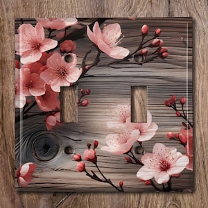 May include: A decorative light switch cover featuring a floral design. The cover has a wooden background with pink cherry blossoms and buds. The cover has two switch openings and a glossy finish.