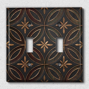 Puede incluir: Una tapa de interruptor de luz doble con un diseño decorativo que presenta un patrón repetitivo de círculos superpuestos y flores estilizadas en tonos marrones, dorados y negros.
