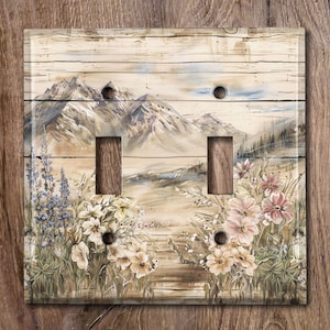 May include: A double light switch cover with a rustic wood background. The image features a mountain scene with wildflowers in the foreground. The switch cover is a light beige color with a distressed wood look.