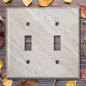 May include: A light beige double toggle light switch cover with a marble pattern. The cover has two toggle switches and two screw holes.