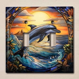 May include: A stained glass style light switch cover featuring a dolphin leaping out of the water at sunset. The dolphin is blue and gray, and the water is blue and green. The sunset is orange and yellow, and the sky is a light blue. The background is a dark brown.