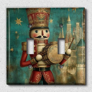 May include: A decorative light switch cover featuring a vintage illustration of a nutcracker soldier playing a drum. The artwork has a teal background with gold stars and a castle in the distance. The nutcracker is dressed in a red and gold uniform.