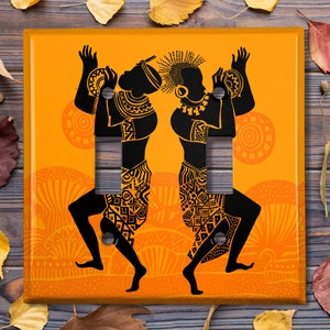 May include: A pair of orange light switch covers with a black silhouette of two figures dancing. The figures are wearing traditional African clothing and holding drums. The background is a pattern of orange and black circles.