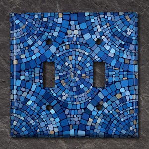 May include: A decorative double light switch cover with a mosaic tile design. The cover features a pattern of blue, light blue, and gray tiles arranged in a circular pattern. The cover has a glossy finish and is mounted on a dark gray surface.