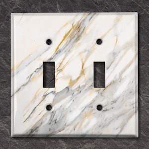 May include: A white light switch cover with a marble pattern featuring gold and grey veins. The cover has two toggle switches and two screw holes.