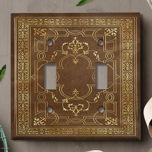 May include: A brown leather-look light switch cover with a gold ornate border and two toggle switches. The cover has a decorative design in gold.