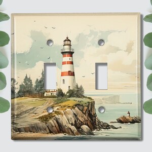 May include: A light switch cover featuring a watercolor painting of a lighthouse on a rocky cliff overlooking the ocean. The lighthouse is white with red stripes and a small, white house at its base. The ocean is a light blue color with white waves.