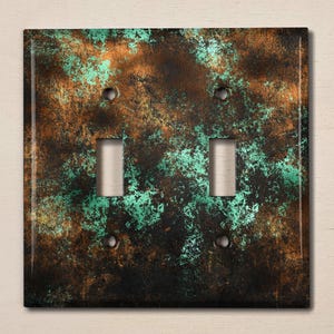 May include: A double light switch cover with a rustic, distressed copper and green finish. The cover has a mottled, aged appearance with a mix of brown, copper, and green tones.