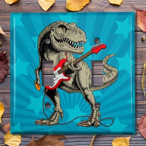 May include: A light blue light switch cover featuring a cartoon dinosaur playing a red electric guitar. The dinosaur is green and has a blue background with white stars.