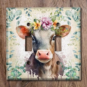 May include: A watercolor painting of a cow with a floral crown on its head. The cow is brown and white with a pink nose and black eyes. The background is a light blue with green leaves.