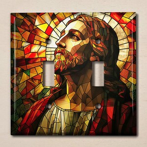 May include: A decorative light switch cover featuring a stained-glass design. The artwork depicts a man with a beard, looking upwards, with a halo. The color palette includes reds, yellows, and oranges, creating a religious theme.
