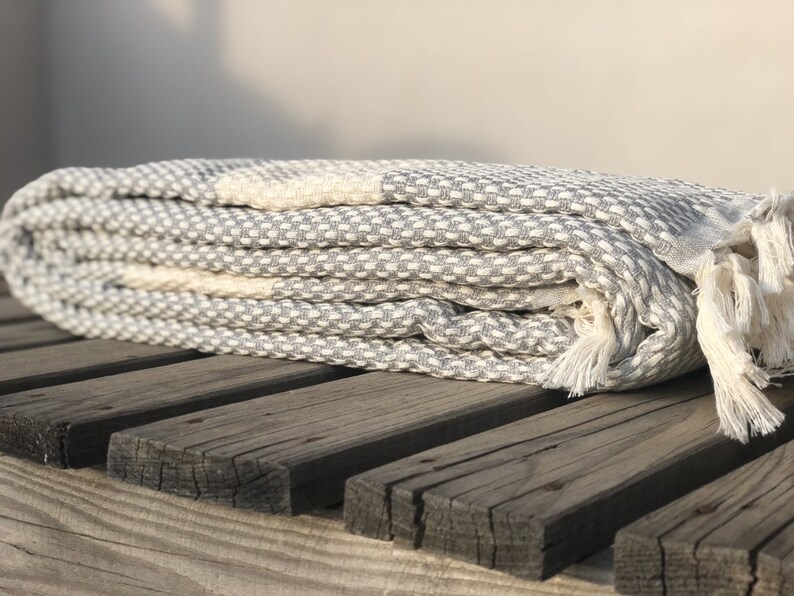 May include: A gray and white waffle weave Turkish towel with fringe. The towel is folded and placed on a wooden surface.