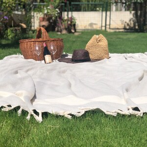 May include: A white picnic blanket with a woven texture and fringe edges. A wicker basket, two wine glasses, a bottle of wine, a straw hat, and a black hat are on the blanket.