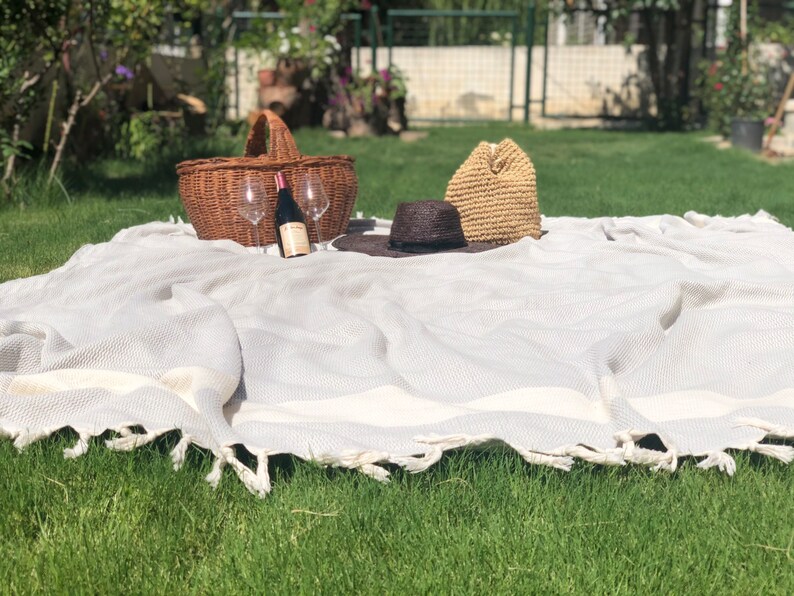 May include: A white picnic blanket with a woven texture and fringe edges. A wicker basket, two wine glasses, a bottle of wine, a straw hat, and a black hat are on the blanket.