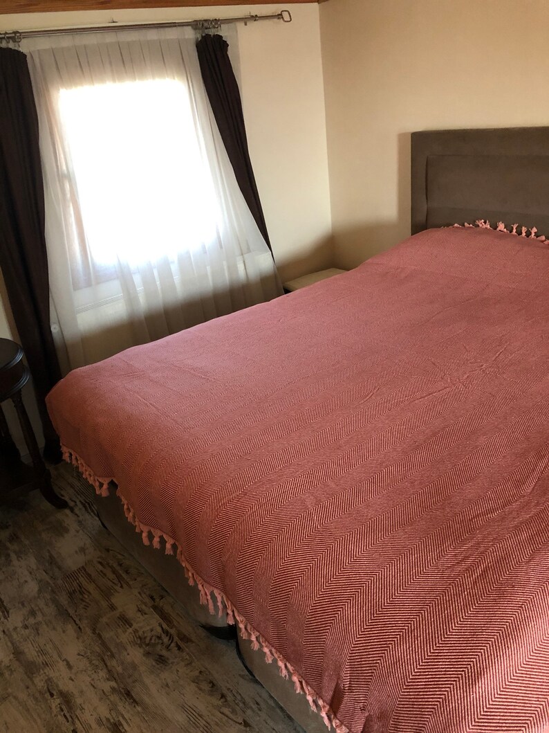 May include: A bedroom with a large bed with a pink and white chevron patterned duvet cover. The bed has a brown headboard and a white nightstand. The room has a window with white curtains and brown drapes.